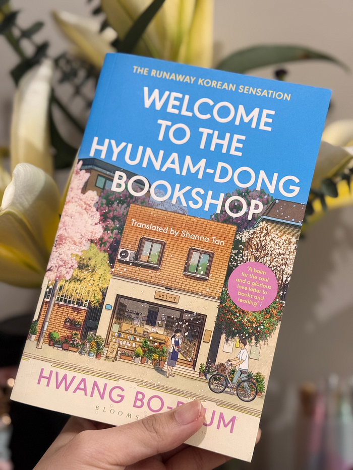 A person holding the book 'Welcome to the Hyunam-dong Bookshop' against a floral background, showcasing its colorful cover and details.